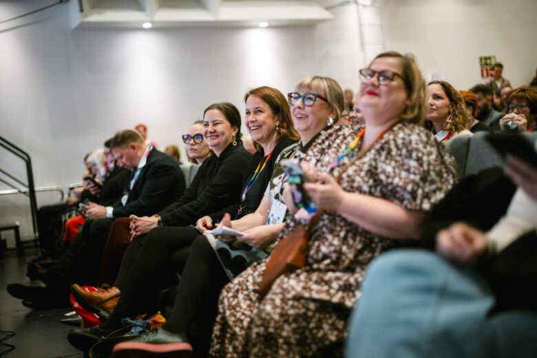 A diverse group of people sit in a conference hall, facing forward. Some are smiling and looking towards the stage, while others are looking at their phones. The lighting is bright, and the background is a plain white wall with some ceiling fixtures visible.