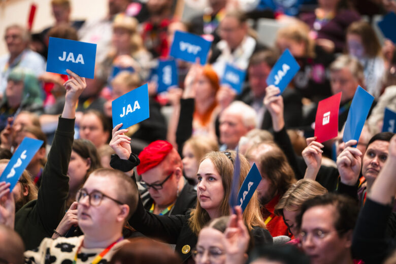 A crowd of people are visible, many holding up blue cards with the word "JAA" in white letters. One person in the foreground holds a red card with a white symbol. The audience is blurred, with focus on the hands and cards.